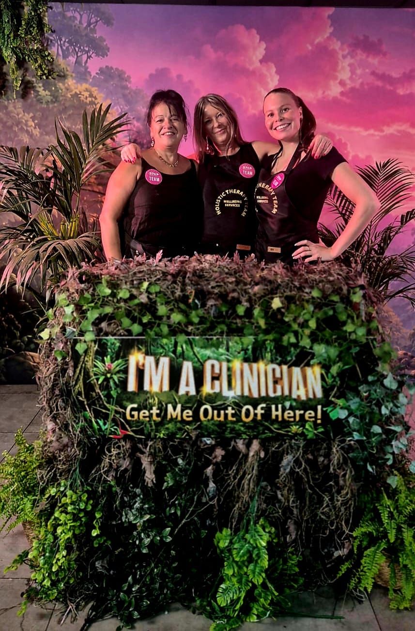Three therapists smiling behind a leafy conference display that reads I'm a Clinician, Get Me Out of Here