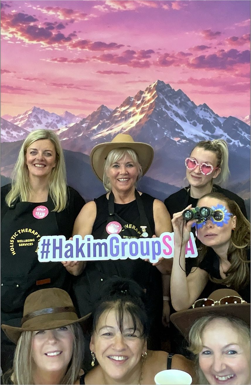 Six of the team in hats and novelty glasses holding a HakimGroupSPA sign in front of a mountain backdrop