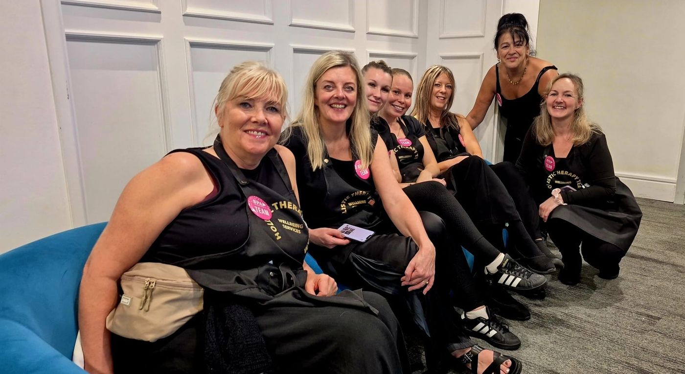 Seven therapists from the Holistic Therapy Team in black aprons, sitting together around a blue sofa