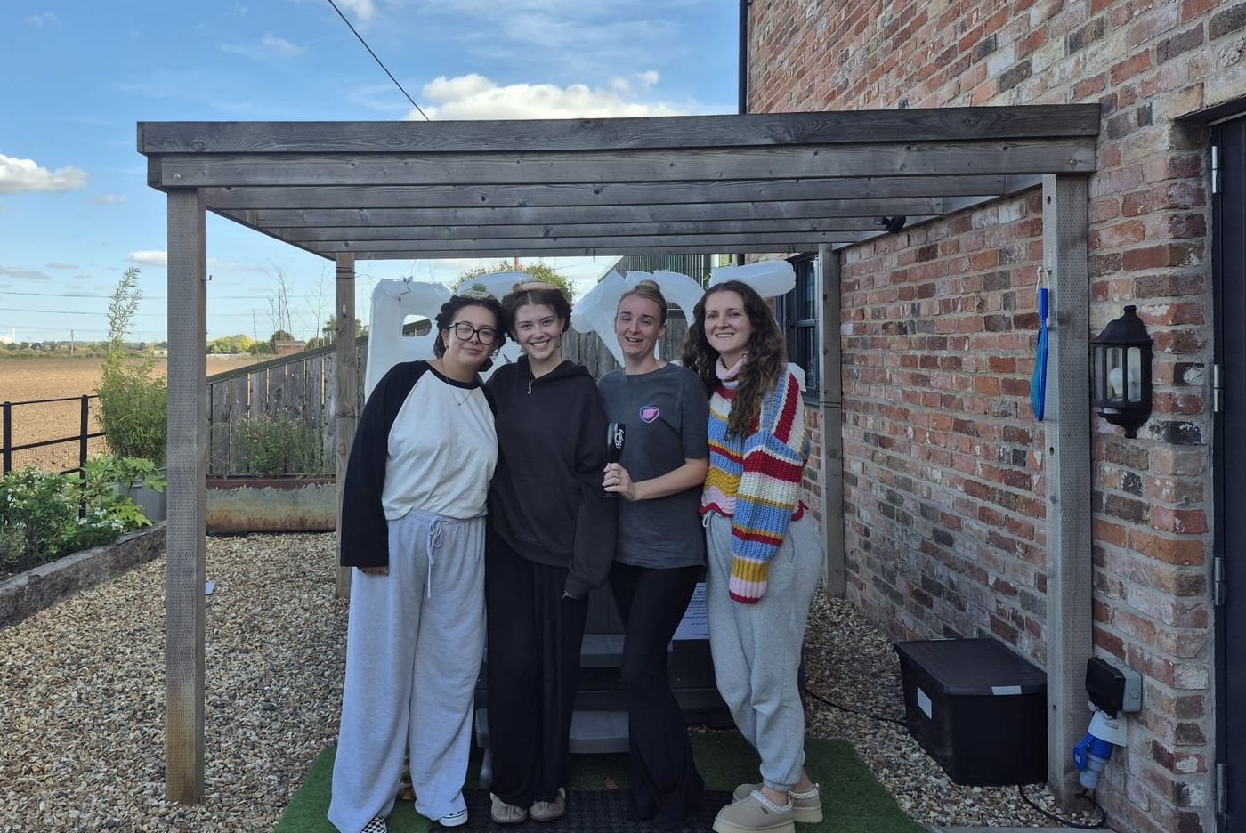 Four of the hen party smiling under a wooden pergola outside the brick barn, with open fields behind
